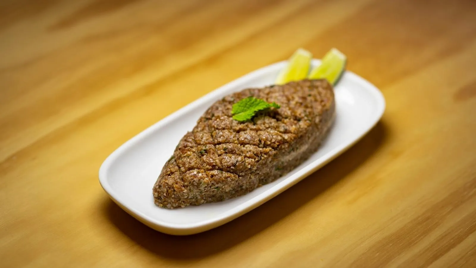 قطعة الكبة النية بيضاوية مزخرفة على طبق أبيض صغير مع ورقة نعناع وشرائح ليمون فوق طاولة خشبية