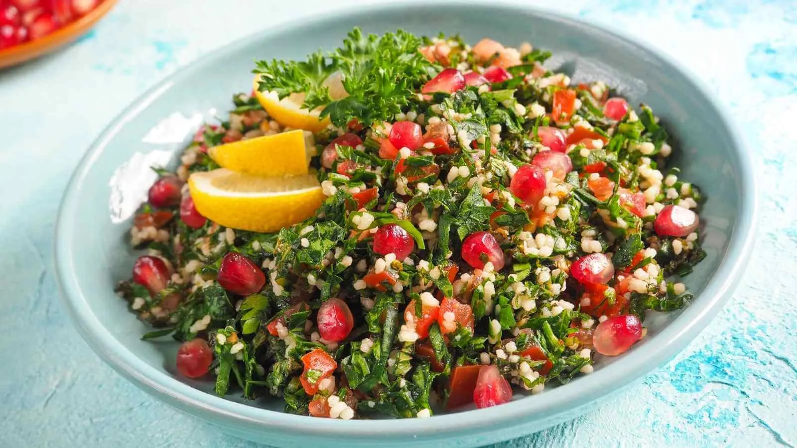 تبولة مع حبّات رمان وشرائح ليمون في طبق أزرق على خلفية فيروزية